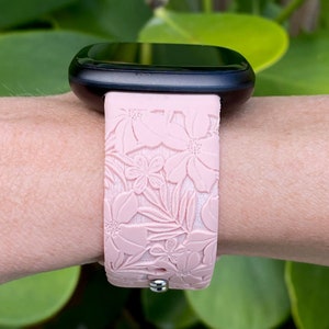 Puede incluir: Una correa de reloj de silicona rosa con un patr&oacute;n floral. La correa se lleva en la mu&ntilde;eca y tiene una hebilla plateada.