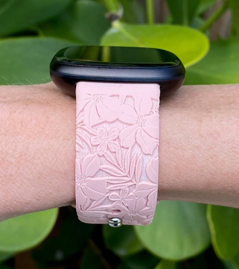 Puede incluir: Una correa de reloj de silicona rosa con un patr&oacute;n floral. La correa se lleva en la mu&ntilde;eca y tiene una hebilla plateada.