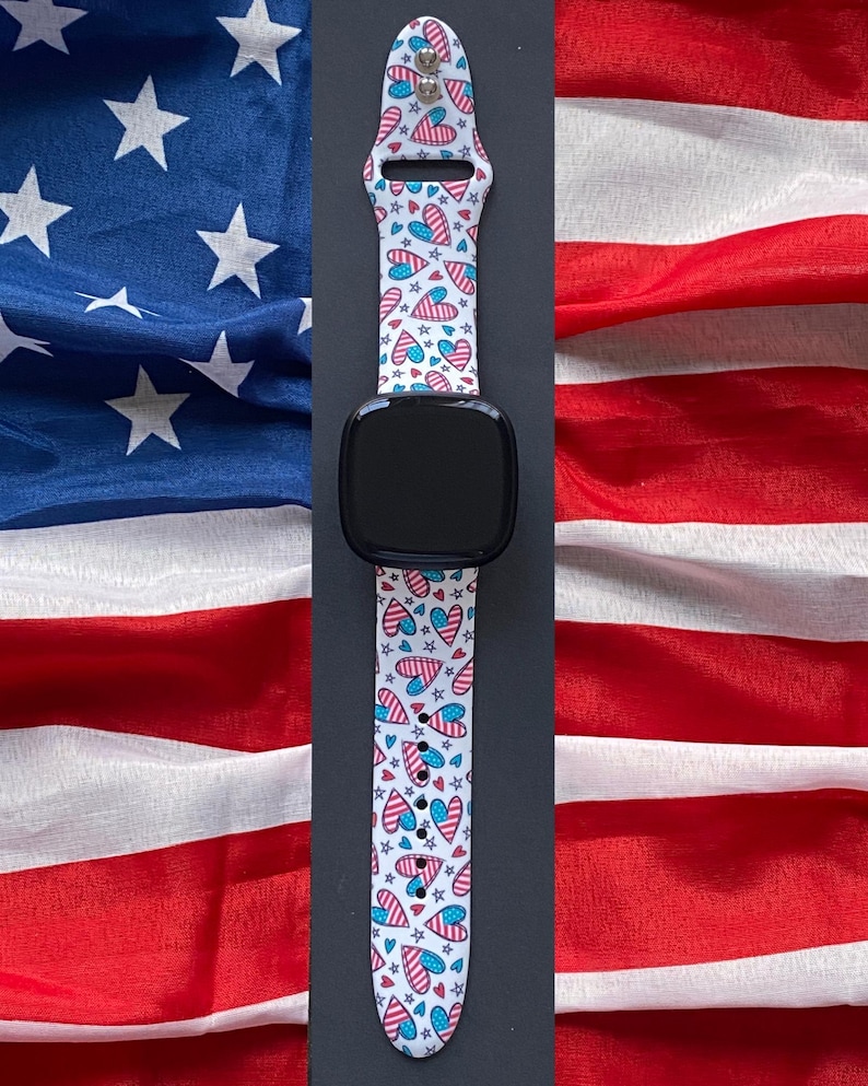 May include: A black smartwatch with a white strap patterned with red, white, and blue hearts and stars. The background features the American flag.