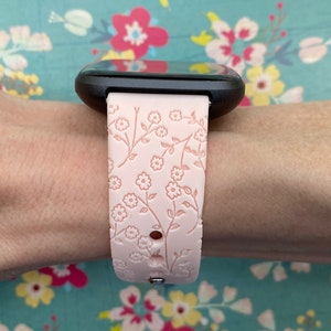 May include: A pink silicone watch band with a floral pattern. The band is designed for a smartwatch with a black face.