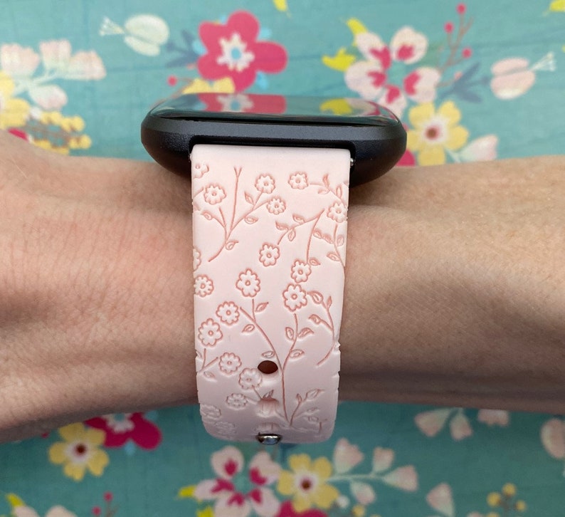 May include: A pink silicone watch band with a floral pattern. The band is designed for a smartwatch with a black face.