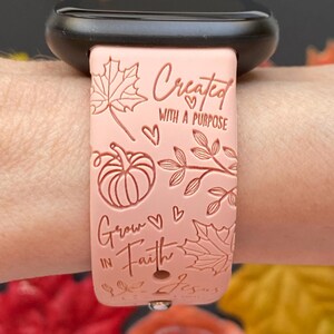 May include: A light pink watch band with the text "Created with a Purpose" and "Grow in Faith". The band features leaf and pumpkin designs. The watch band is attached to a black watch.