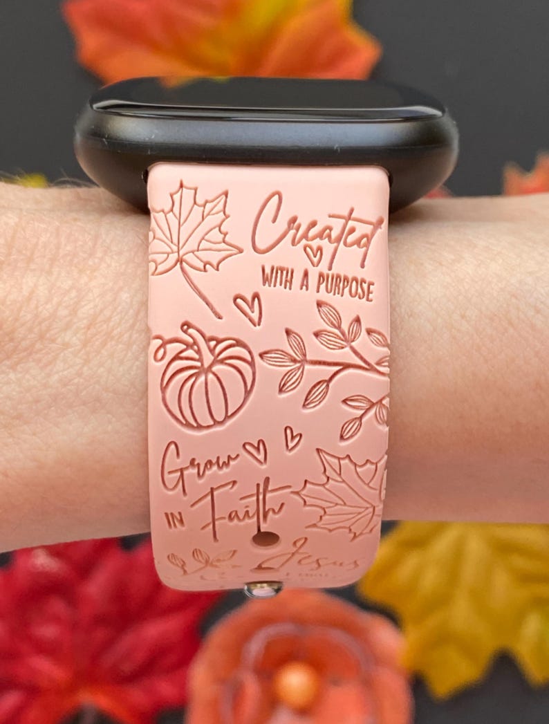 May include: A light pink watch band with the text "Created with a Purpose" and "Grow in Faith". The band features leaf and pumpkin designs. The watch band is attached to a black watch.