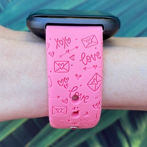 May include: Pink silicone watch band with a pattern of hearts, arrows, and envelopes with the words "XOXO" and "love" printed on it.