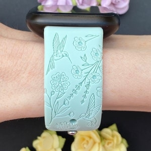 May include: A light blue silicone watch strap with a floral and hummingbird design. The strap is shown on a wrist.