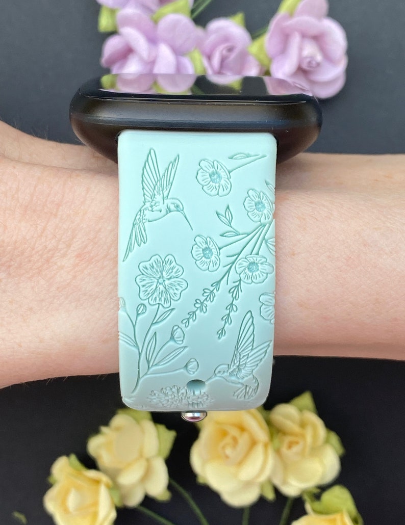 May include: A light blue silicone watch strap with a floral and hummingbird design. The strap is shown on a wrist.