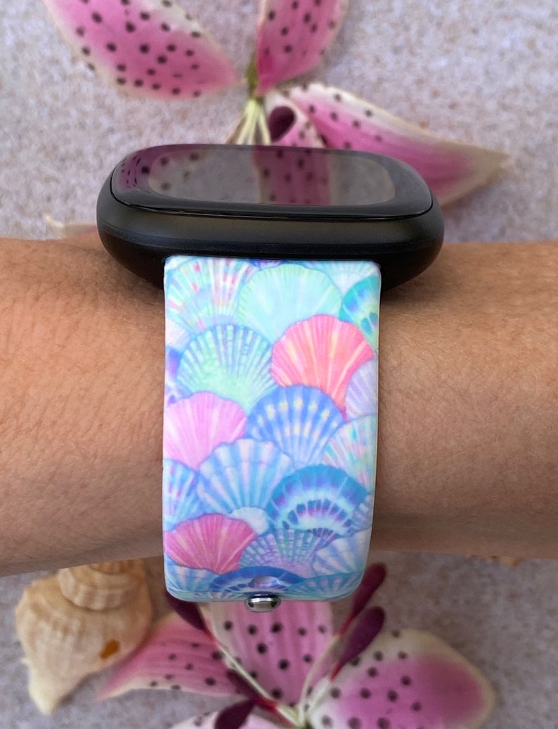 K&ouml;nnte beinhalten: Eine Smartwatch mit einem schwarzen Armband mit einem bunten Muschelmuster. Das Armband ist aus Silikon und hat einen wei&szlig;en Hintergrund mit blauen, rosa und gr&uuml;nen Muscheln.