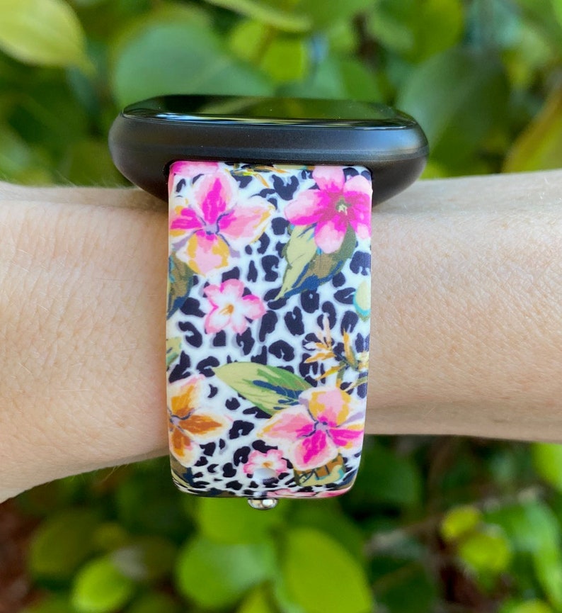 May include: A black smartwatch with a floral and leopard print silicone strap. The strap features pink, white, and yellow flowers on a black and white leopard print background.