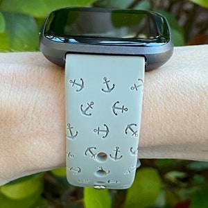 May include: A gray fitness tracker with a gray band featuring a repeating pattern of small anchors. The band is worn on a person's wrist.