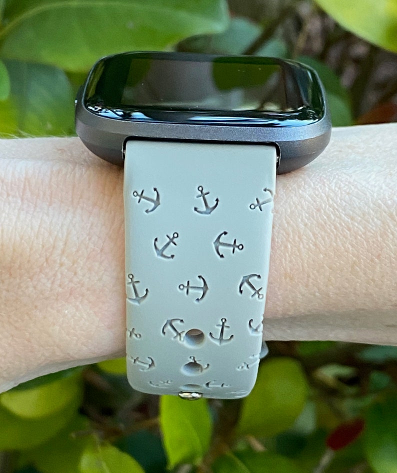 May include: A gray fitness tracker with a gray band featuring a repeating pattern of small anchors. The band is worn on a person's wrist.