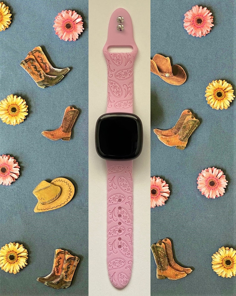 May include: A pink smartwatch band with a paisley pattern. The band is shown on a white background with various decorative items, including pink flowers, cowboy hats, and cowboy boots.