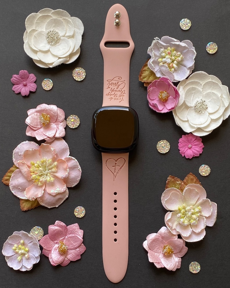 May include: A pink silicone watch band with a black watch face. The band has the words "Love always,  strength  within,  grace  around" engraved on it. The watch face has a heart with a cross inside it.