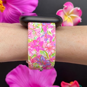 K&ouml;nnte beinhalten: Eine schwarze Apple Watch mit einem wei&szlig;en Armband mit Blumenmuster. Das Armband zeigt rosa, lila und gr&uuml;ne Blumen auf wei&szlig;em Hintergrund.