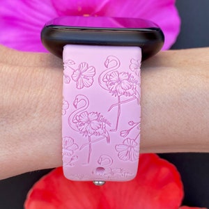 May include: A light pink watch band with a flamingo and floral design. The band is attached to a black smartwatch. The background includes a pink and red flower.