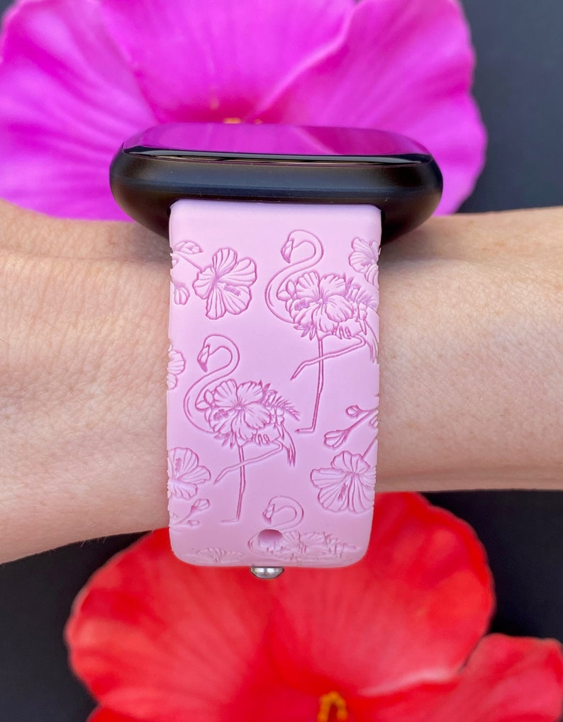 May include: A light pink watch band with a flamingo and floral design. The band is attached to a black smartwatch. The background includes a pink and red flower.
