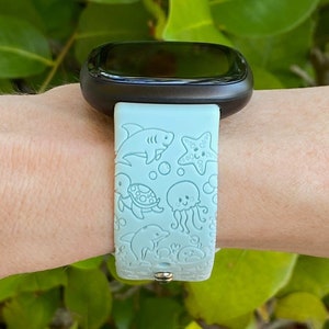 May include: A light blue smartwatch band with a sea-themed design. The band features a shark, starfish, turtle, jellyfish, and dolphin.