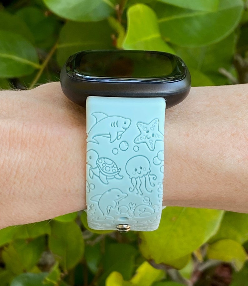 May include: A light blue smartwatch band with a sea-themed design. The band features a shark, starfish, turtle, jellyfish, and dolphin.