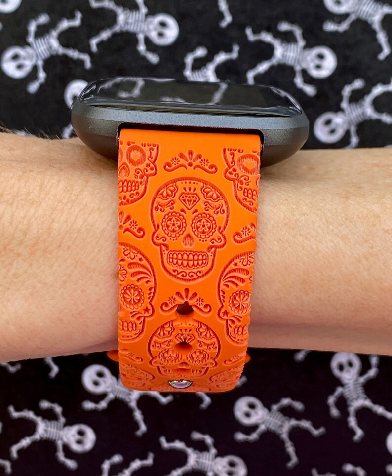 K&ouml;nnte beinhalten: Orangefarbenes Silikon-Armband mit Totenkopf-Design. Das Armband wird am Handgelenk getragen.