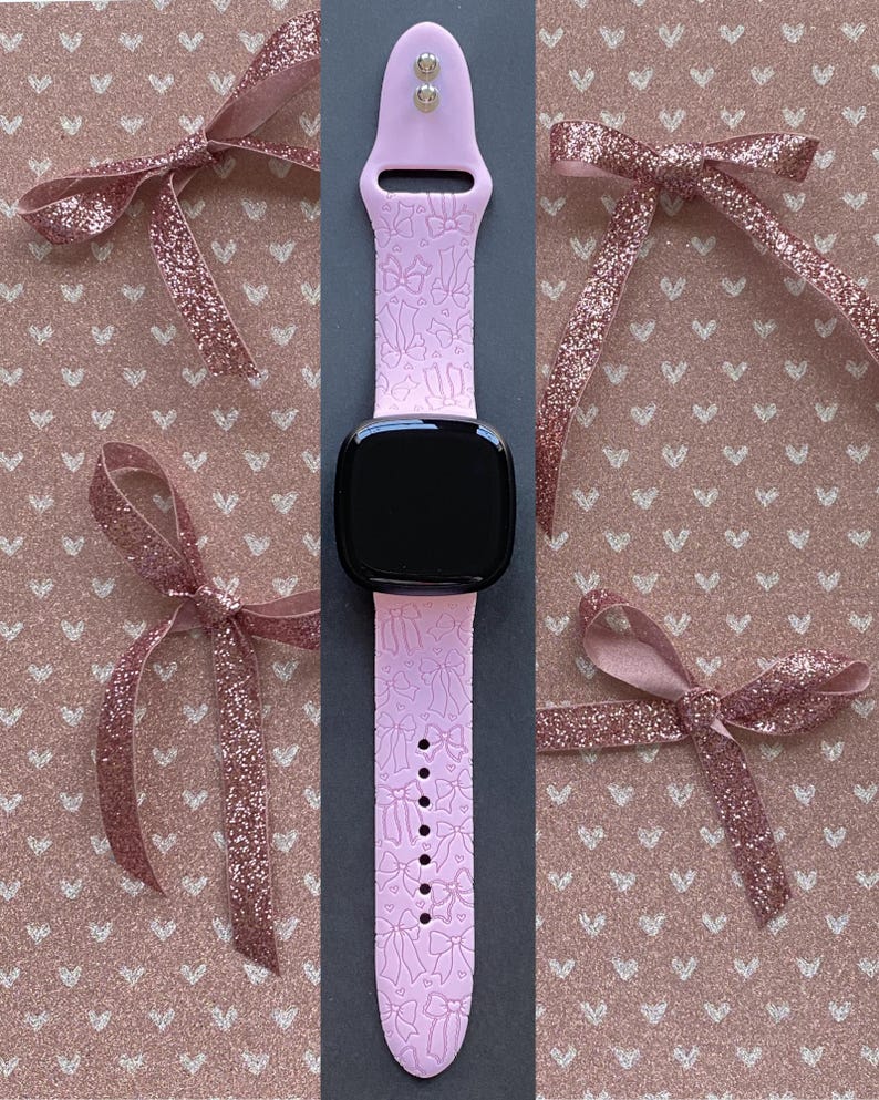 May include: A light pink smartwatch band with a pattern of bows and ballet shoes. The band is displayed with a black smartwatch face and surrounded by pink glitter bows on a pink background.