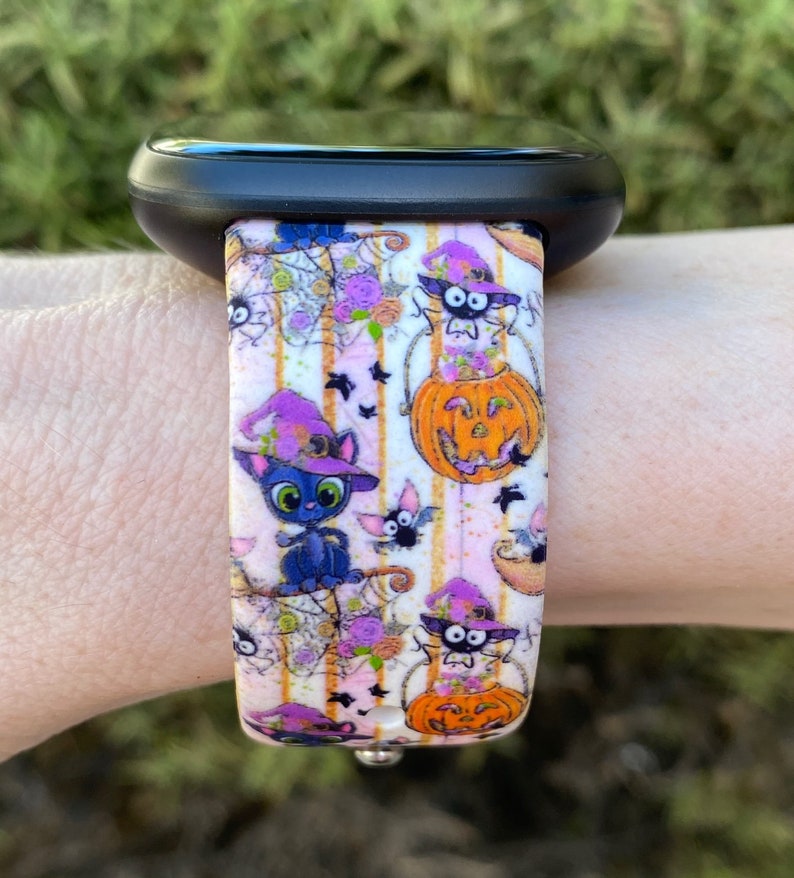 以下が含まれることがあります： ハロウィンをテーマにした白い腕時計のバンド。バンドには、魔女の帽子をかぶった黒い猫、カボチャ、コウモリがピンクと白のストライプの背景に描かれています。