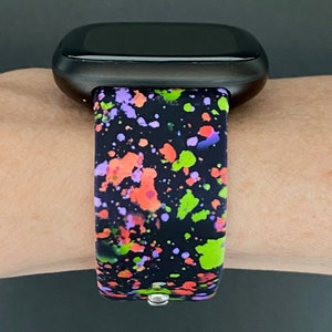 Puede incluir: Un reloj inteligente negro con una banda de silicona de pintura salpicada de colores. La banda tiene salpicaduras de pintura verde, naranja, rosa y p&uacute;rpura sobre un fondo negro.