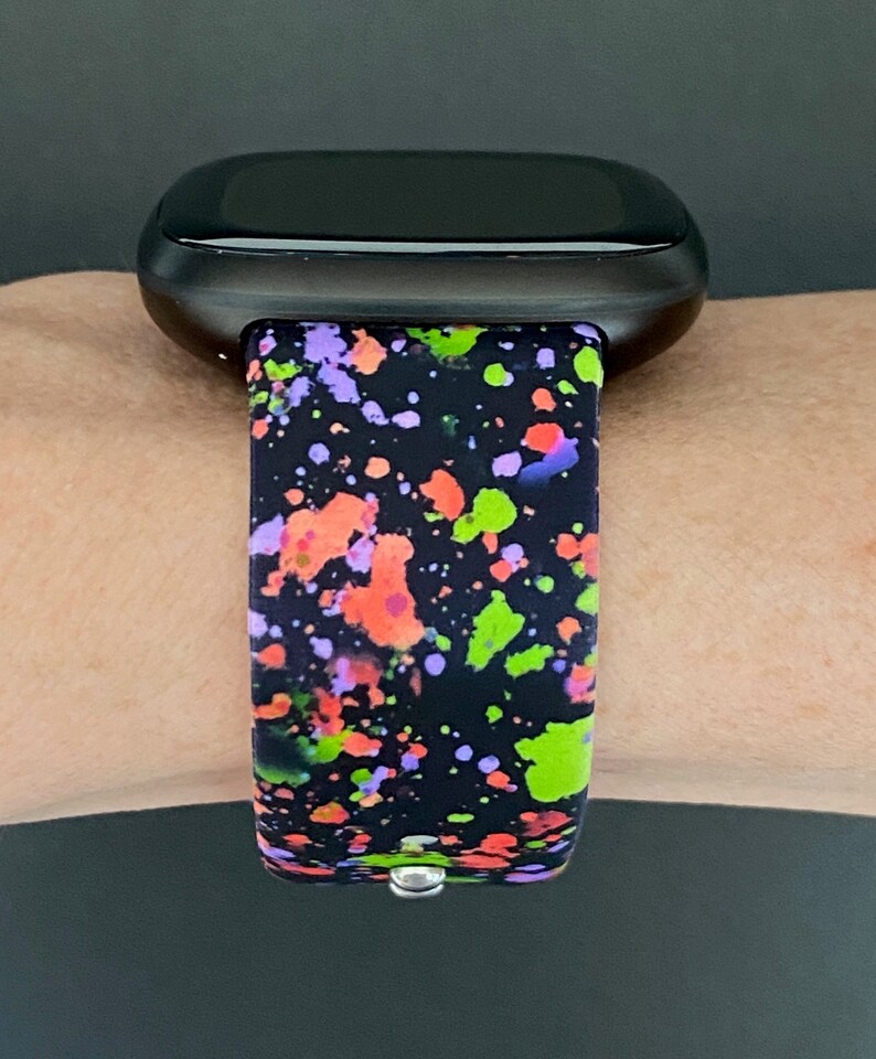 Puede incluir: Un reloj inteligente negro con una banda de silicona de pintura salpicada de colores. La banda tiene salpicaduras de pintura verde, naranja, rosa y p&uacute;rpura sobre un fondo negro.