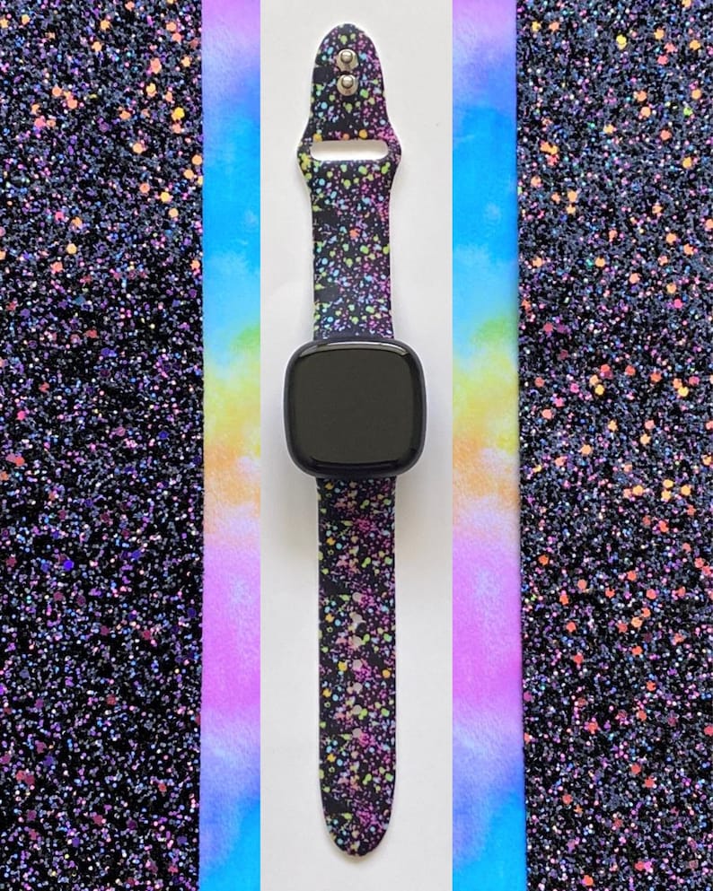May include: A black smartwatch band with a colorful splatter pattern. The band is made of silicone and has a silver buckle. The band is shown against a background of iridescent glitter.