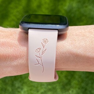 May include: A smartwatch with a gray case and a light pink band. The band features a floral design. The watch is on a wrist against a blurred green background.
