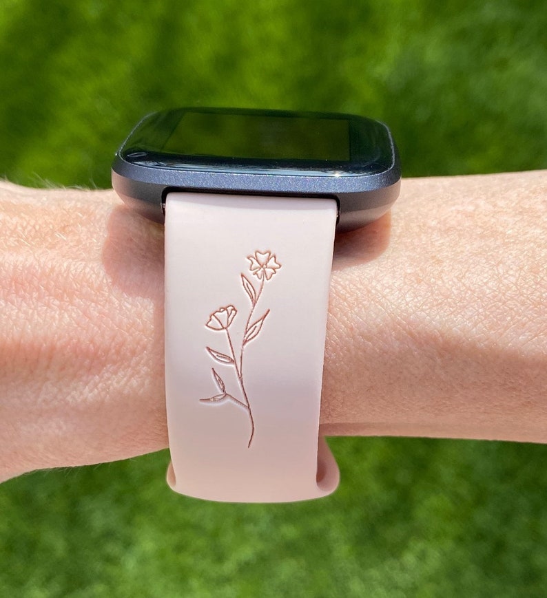 May include: A smartwatch with a gray case and a light pink band. The band features a floral design. The watch is on a wrist against a blurred green background.