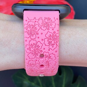 May include: Pink silicone watch band with a hibiscus flower pattern. The band is designed for a smartwatch and has a black watch face.