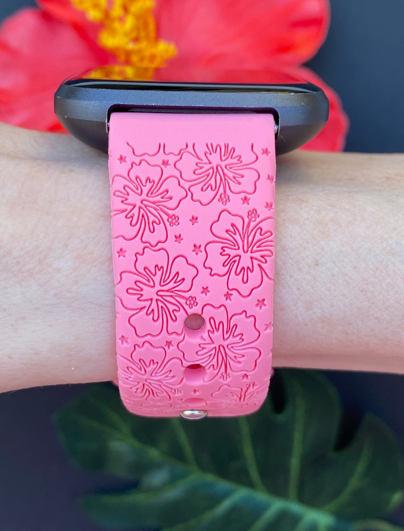 May include: Pink silicone watch band with a hibiscus flower pattern. The band is designed for a smartwatch and has a black watch face.