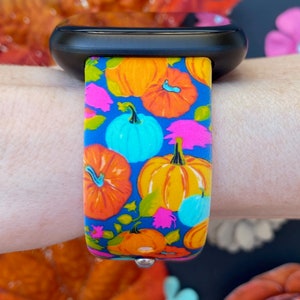 May include: A black smartwatch with a colorful pumpkin and floral patterned band. The band is blue with orange, yellow, pink, and green pumpkins and flowers.