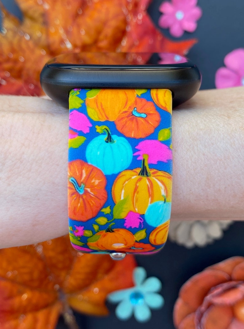 May include: A black smartwatch with a colorful pumpkin and floral patterned band. The band is blue with orange, yellow, pink, and green pumpkins and flowers.
