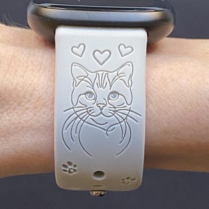 May include: A light gray smartwatch band featuring a cat design with hearts and paw prints. The cat's face is centered, with three hearts above and paw prints below. The watch face is black.