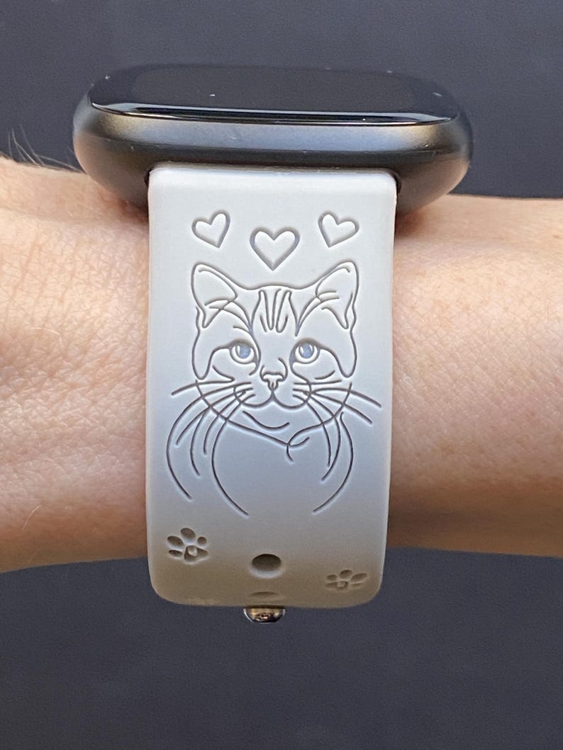 May include: A light gray smartwatch band featuring a cat design with hearts and paw prints. The cat's face is centered, with three hearts above and paw prints below. The watch face is black.
