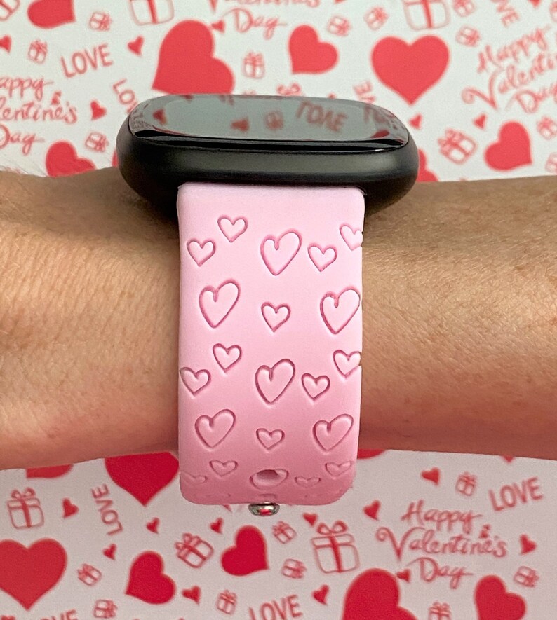 May include: A pink silicone watch strap with a heart pattern. The strap is on a person's wrist.