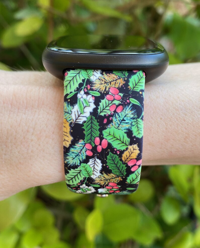 May include: A black smartwatch with a green, red and gold floral patterned band. The band has a repeating pattern of leaves, berries and flowers.