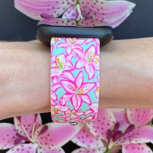 Peut inclure: Une montre connect&eacute;e noire avec un bracelet &agrave; motif floral rose et vert. Le bracelet pr&eacute;sente un motif r&eacute;p&eacute;t&eacute; de lys roses sur un fond vert clair.