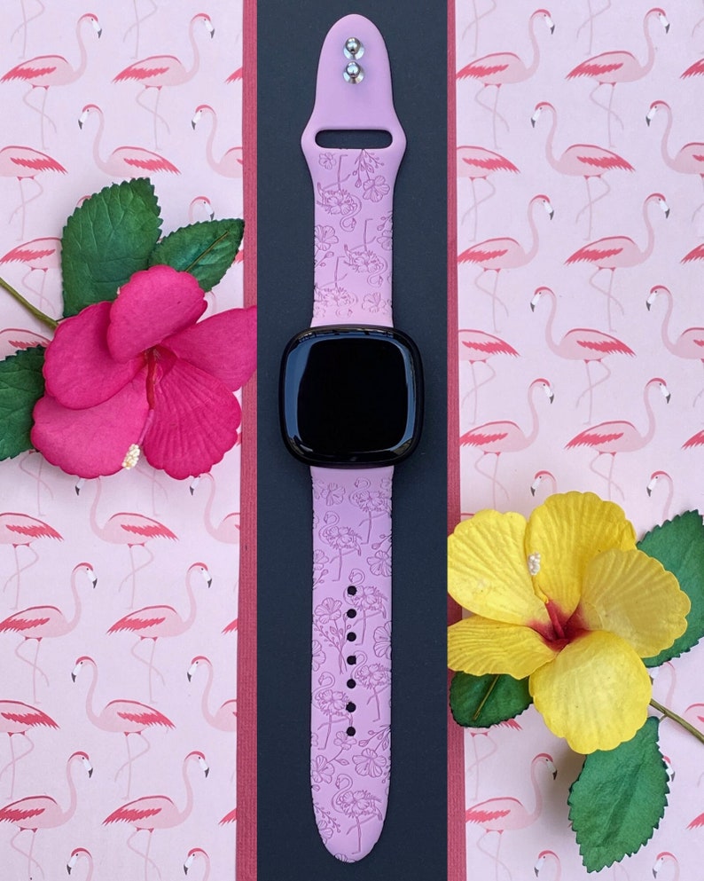 May include: A light purple smartwatch band with a floral pattern, set against a pink flamingo-themed background. The band is paired with a black smartwatch face. Decorative pink and yellow flowers with green leaves are placed around the band.