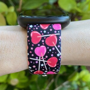 K&ouml;nnte beinhalten: Eine schwarze Smartwatch mit einem pink-schwarzen Silikonarmband mit Herz-Lollipop-Druck. Das Armband hat wei&szlig;e Sterne auf schwarzem Hintergrund.