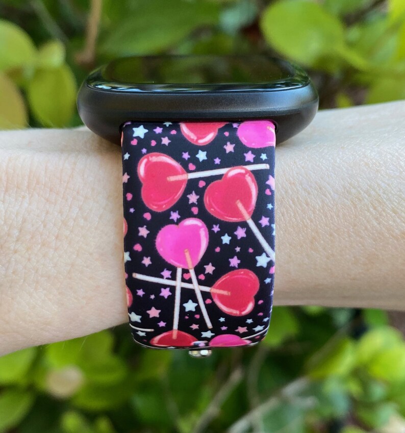 K&ouml;nnte beinhalten: Eine schwarze Smartwatch mit einem pink-schwarzen Silikonarmband mit Herz-Lollipop-Druck. Das Armband hat wei&szlig;e Sterne auf schwarzem Hintergrund.