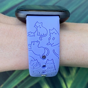 Pode incluir: Uma pulseira de rel&oacute;gio de silicone roxo com um padr&atilde;o de gatos de contorno branco. A pulseira &eacute; projetada para um rastreador de fitness.