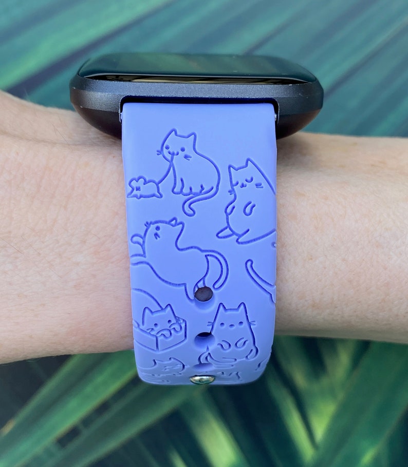 Pode incluir: Uma pulseira de rel&oacute;gio de silicone roxo com um padr&atilde;o de gatos de contorno branco. A pulseira &eacute; projetada para um rastreador de fitness.