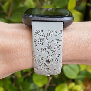 May include: Gray smartwatch band with a pattern of smiley faces, dog bones, and flowers. The band is attached to a black smartwatch. The background is blurred green foliage.