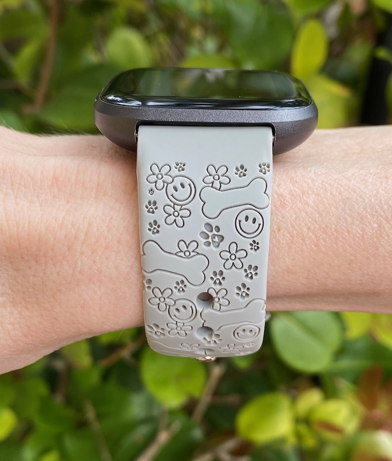 May include: Gray smartwatch band with a pattern of smiley faces, dog bones, and flowers. The band is attached to a black smartwatch. The background is blurred green foliage.