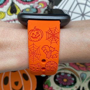 May include: Orange Halloween-themed smartwatch band with jack-o'-lanterns, spiderwebs, a bat, and a skull design. The band is made of a flexible material and is attached to a black smartwatch.