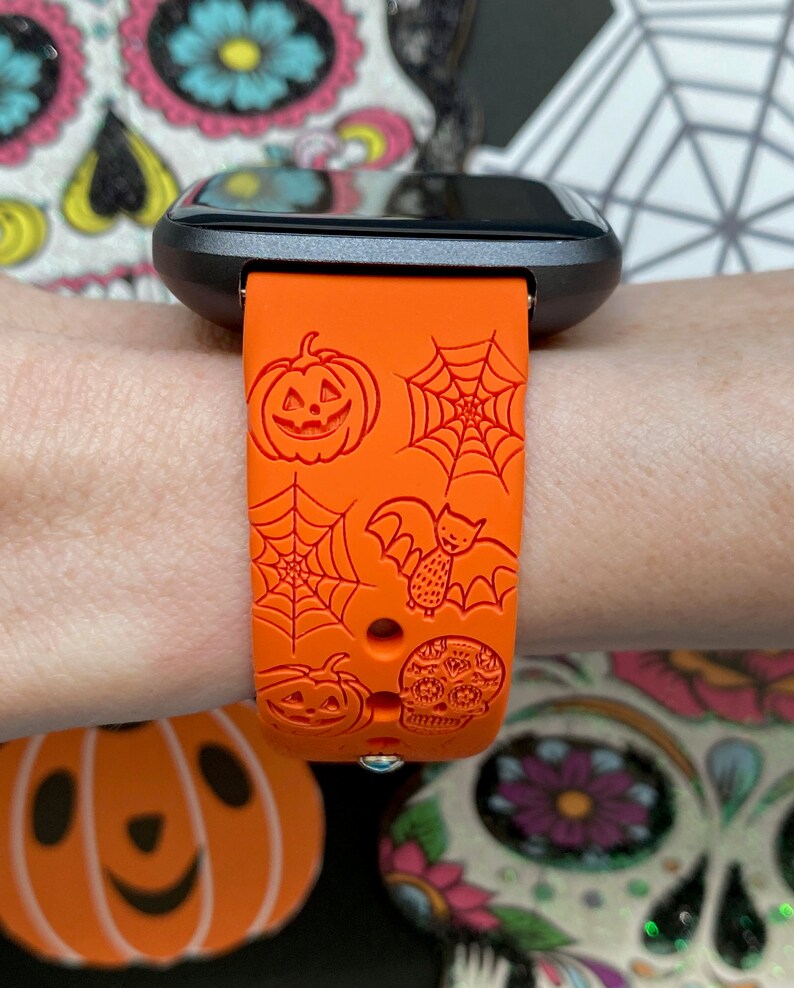 May include: Orange Halloween-themed smartwatch band with jack-o'-lanterns, spiderwebs, a bat, and a skull design. The band is made of a flexible material and is attached to a black smartwatch.