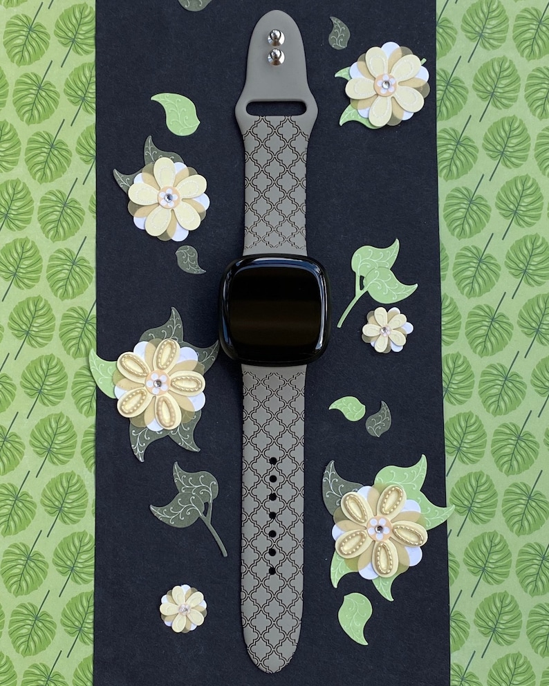K&ouml;nnte beinhalten: Ein graues Armband mit Muster und schwarzem Zifferblatt. Das Armband hat ein geometrisches Muster und ist vor einem schwarzen Hintergrund mit Papierblumen abgebildet.