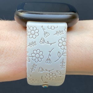 K&ouml;nnte beinhalten: Ein wei&szlig;es Silikon-Armband mit Blumenmuster. Das Armband wird am Handgelenk getragen und hat ein schwarzes Zifferblatt.