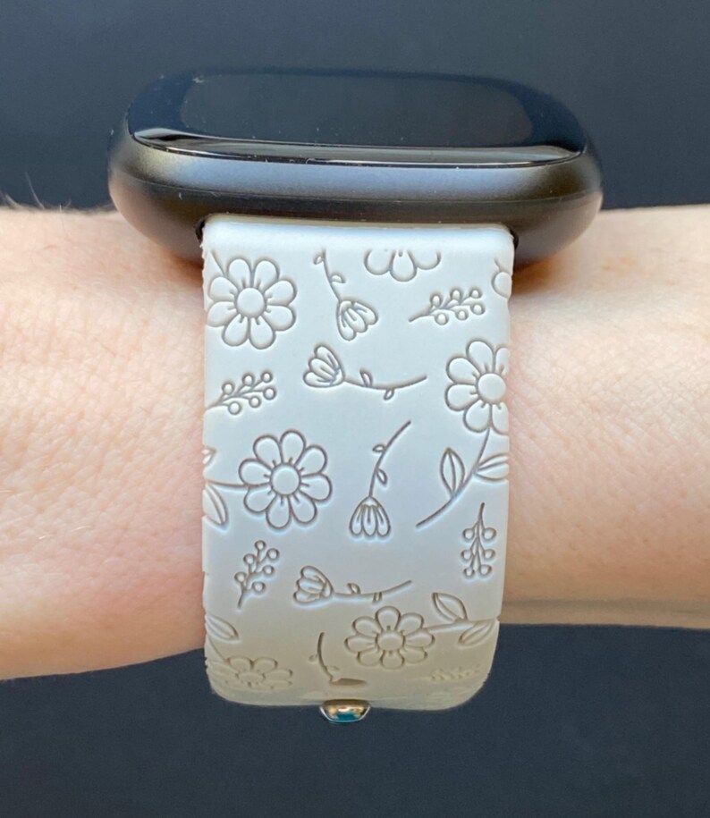 K&ouml;nnte beinhalten: Ein wei&szlig;es Silikon-Armband mit Blumenmuster. Das Armband wird am Handgelenk getragen und hat ein schwarzes Zifferblatt.